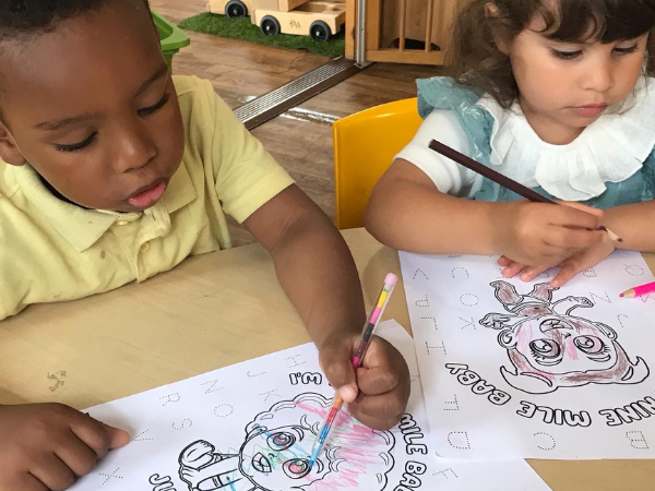 A little black boy and white girl sit next to each other colouring Nine Mile Babies activity sheets of the Nine Mile Babies characters Juls and Prinny Pie at an author visit . The black boy wears a yellow shirt, and the white girl has on a blue dress. The image showcases the focus on providing inclusive and educational experiences for children, highlighting activities for all kids, to develop multicultural diversity by identifying a child’s skin tone.