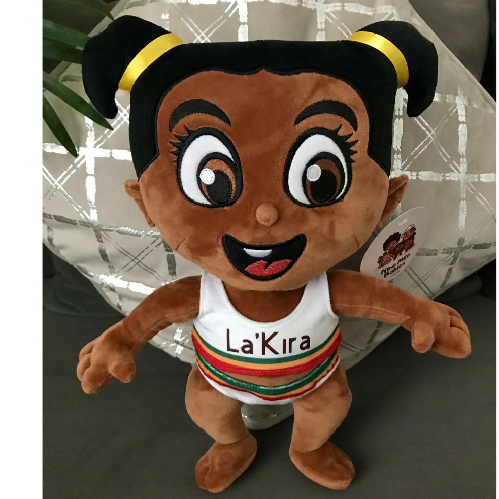 A Nine Mile Babies ‘Prinny Pie’ plush toy doll with a dark skin tone and cheerful expression is standing against a patterned cushion. The doll has large, expressive eyes, pigtails styled with yellow hair bands, and is wearing a white top and bottoms with red, green and yellow stripes on it, personalised with the name "La'Kira" on it. You can personalise your plush toy doll with your own name on your doll. 